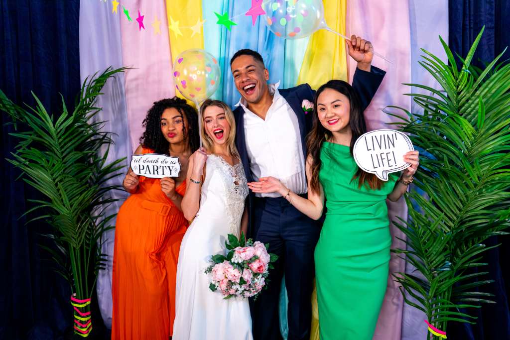 A group of party guests in formal wear stand in front of a colorful backdrop and hold photo props as they pose for a photo